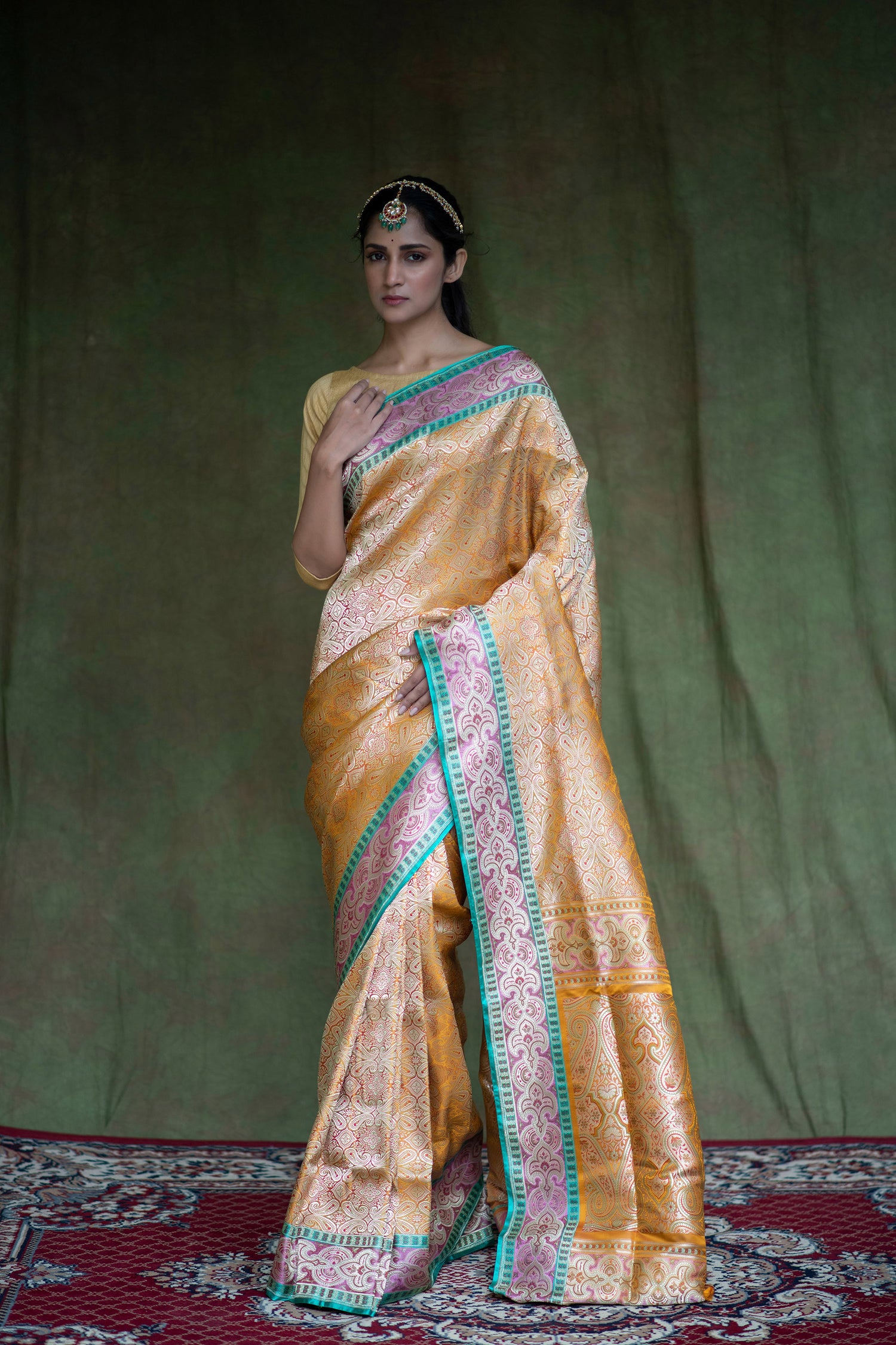 A woman poses gracefully in a mustard yellow Banarasi saree with geometric patterns, floral detailing, and a rich green-gold border.