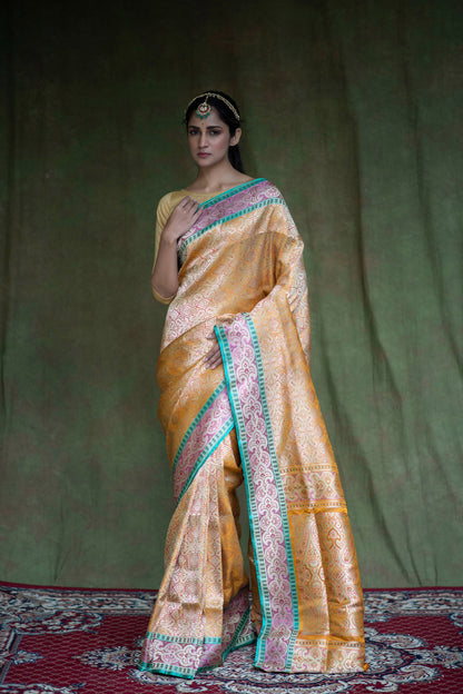 A woman poses gracefully in a mustard yellow Banarasi saree with geometric patterns, floral detailing, and a rich green-gold border.